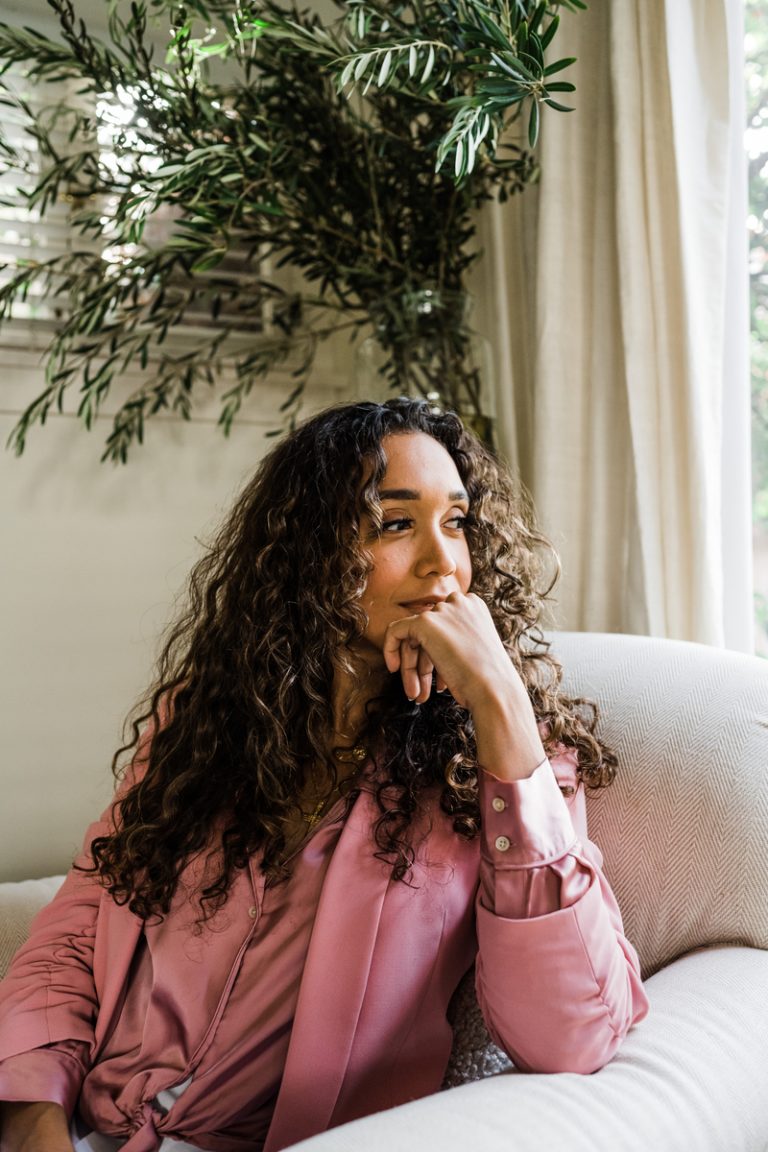 A curly woman in a pink blouse and blazer sits by a sunny window. 
