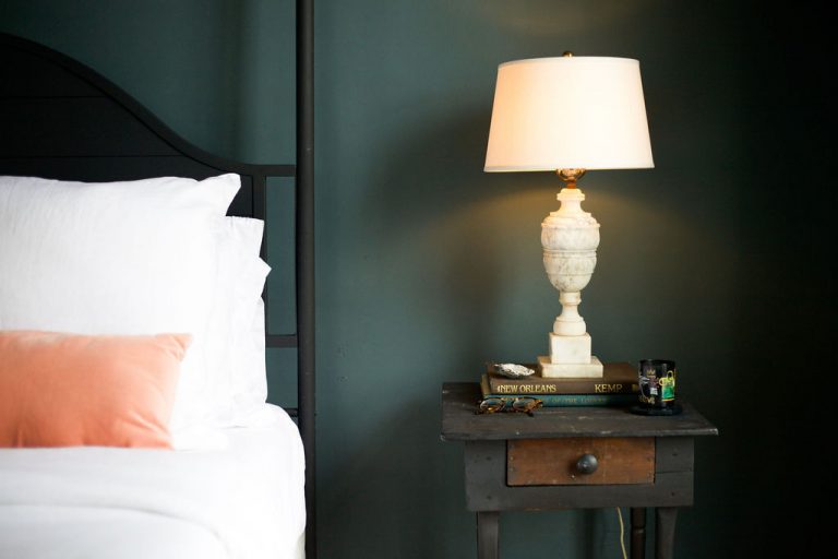Dark green bedroom with white sheets and antique nightstand with white table lamp.