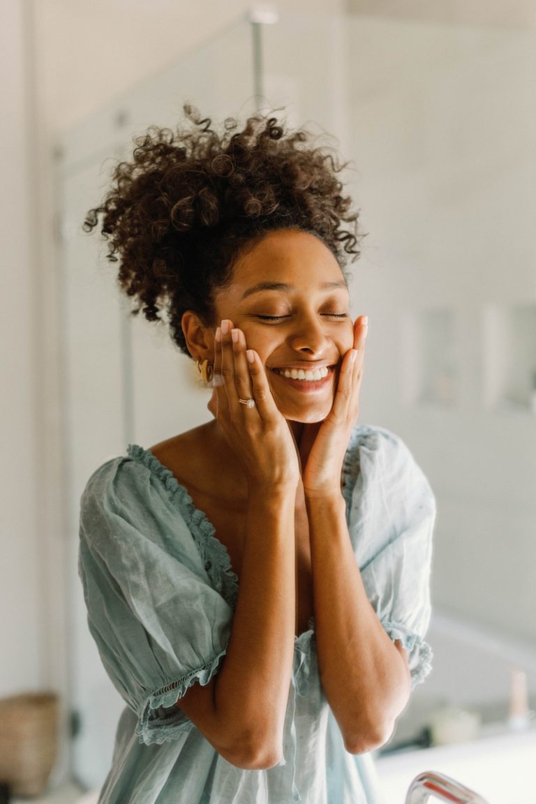 Black woman wearing blue nightgown smiling while washing face in bathroom.