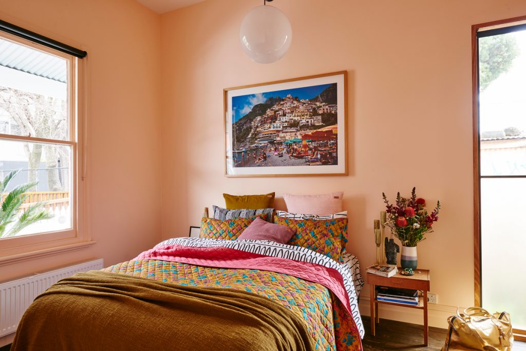 Peach colored bedroom with vibrant orange and pink sheets, beach print hung on wall, and small nightstand with flowers.