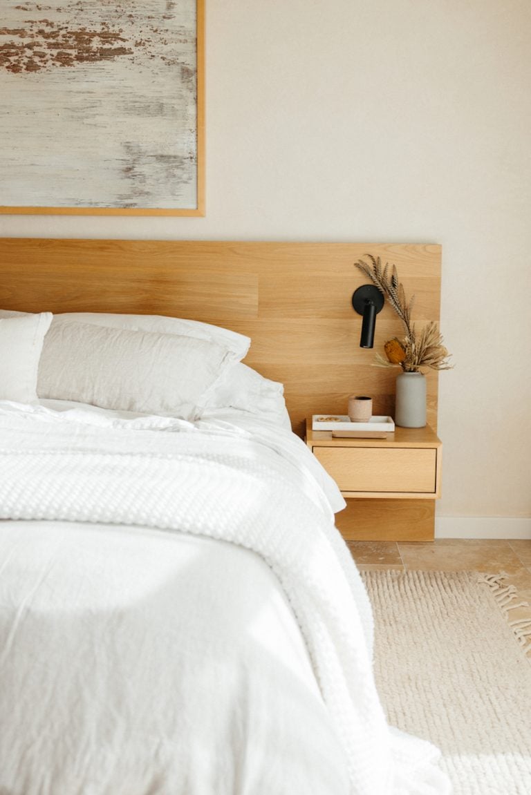 Amplio dormitorio blanco con sábanas blancas, cabecero de madera natural y mesita de noche minimalista con jarrón y flores secas.