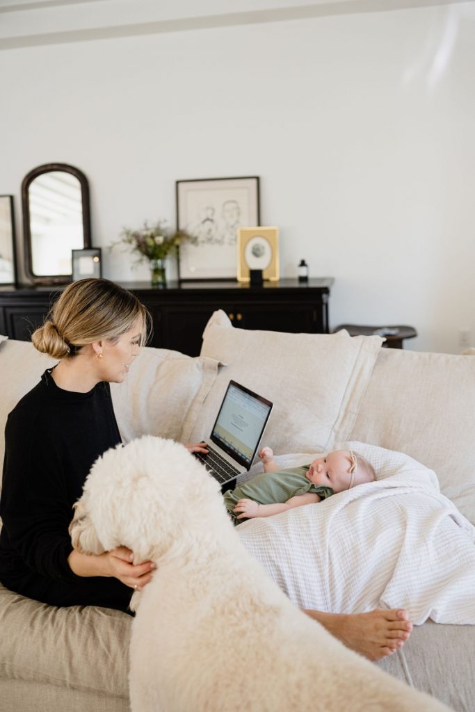 Blonde woman working on laptop on white couch with baby while petting white fluffy dog.