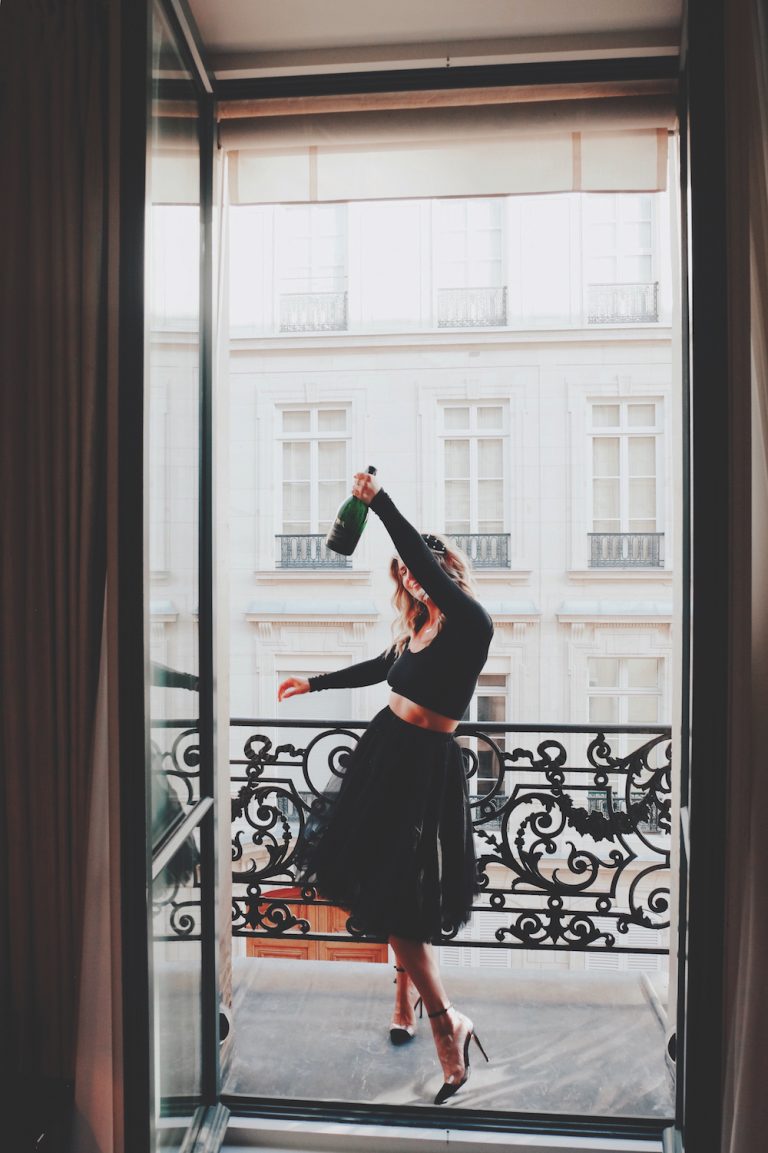 Anna Kloots vestindo saia preta e camisa dançando na varanda em Paris segurando champanhe.