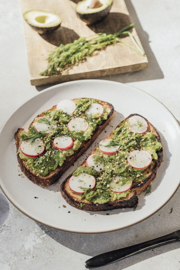 Two slices avocado toast with sliced radishes and herbs on white plate.