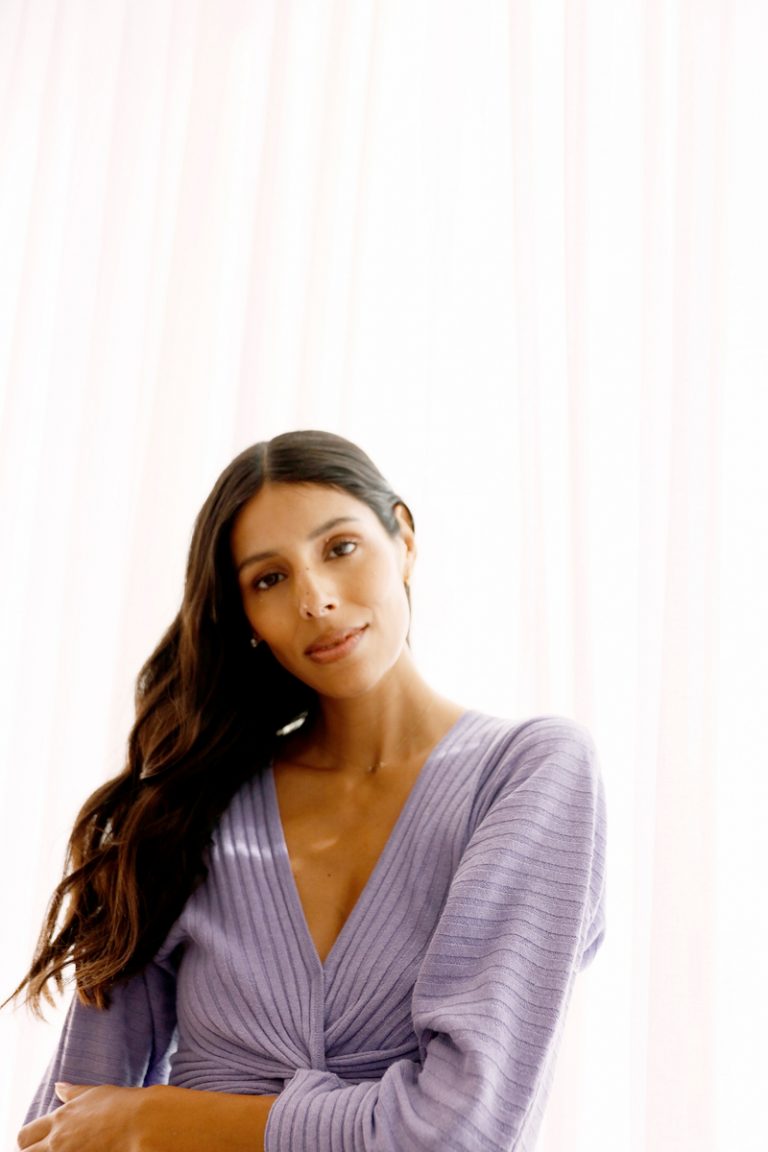 Brunette woman with long wavy hair wearing lilac sweater dress smiling.