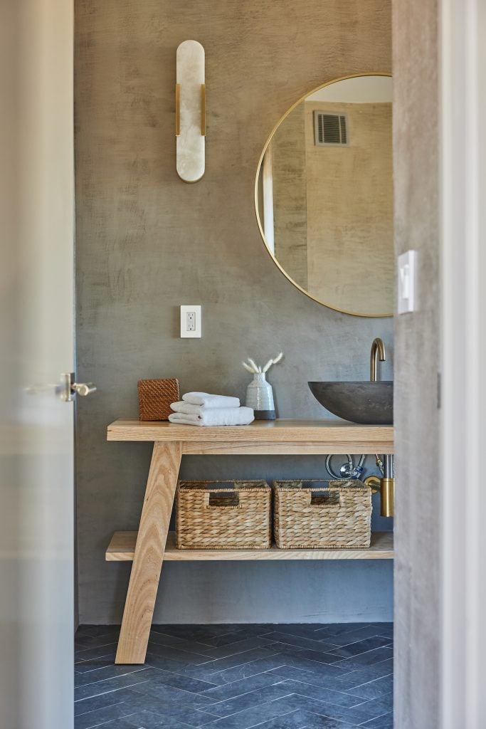 Baño amplio y moderno con paredes de concreto, espejo circular, tocador de madera y accesorios de baño.