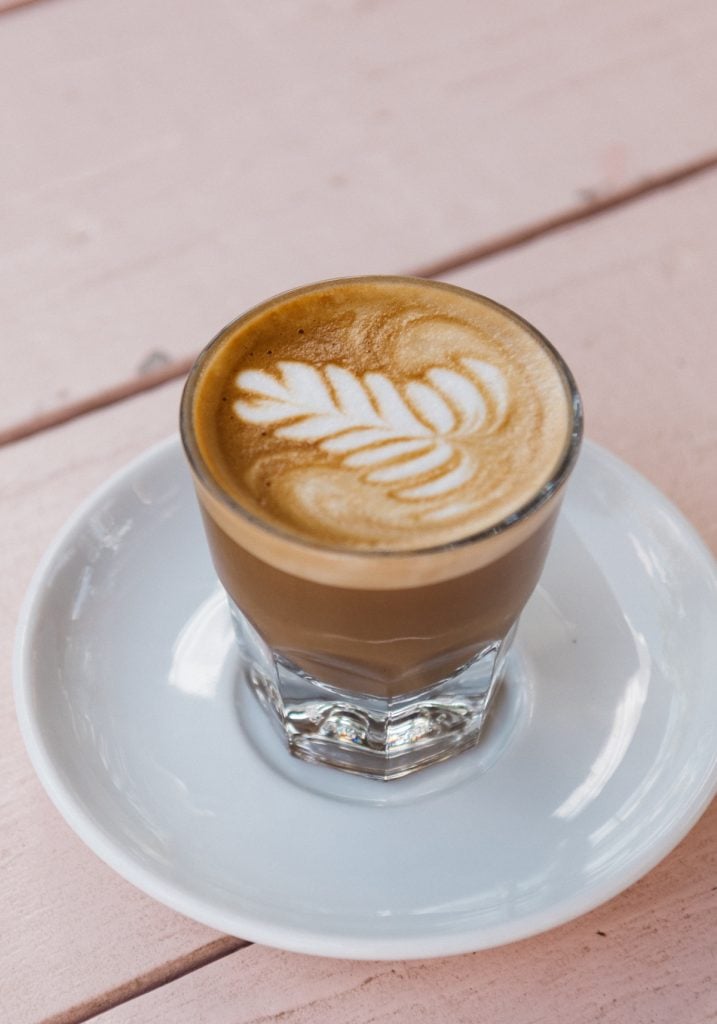 Close-up of latte in glass cup on white plate on light pink picnic table.