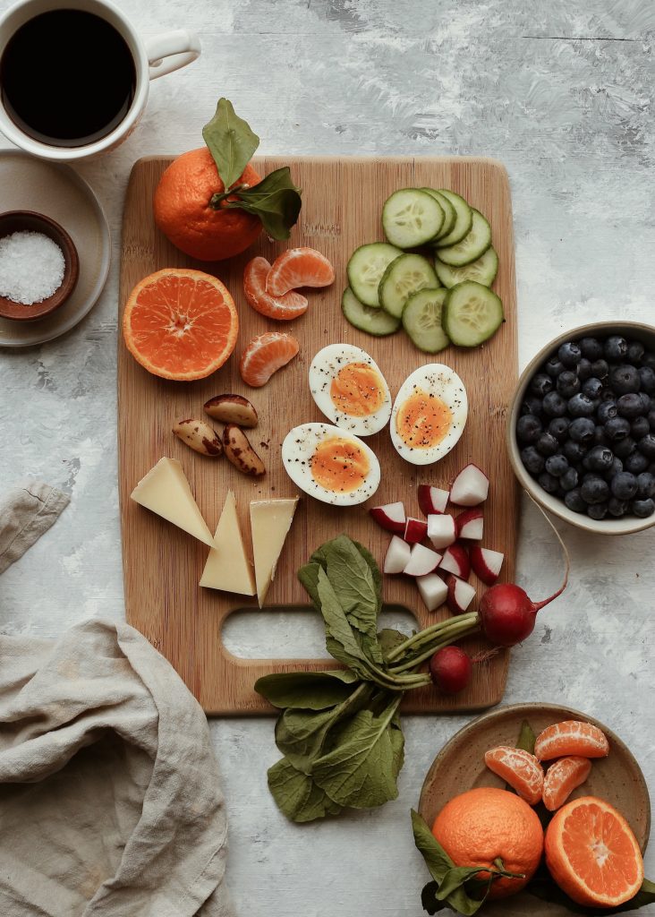 Cutting board on marble countertop with clementines, cucumber slices, Brazil nuts, hardboiled eggs, cheese, radishes, and a bowl of blueberries.