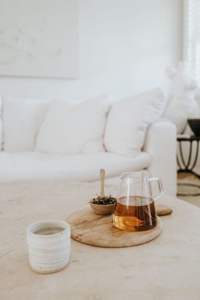 Mesa de centro de mármol con taza de café de gres minimalista, tabla de servir de madera, jarra de té de vidrio frente a un sofá blanco.