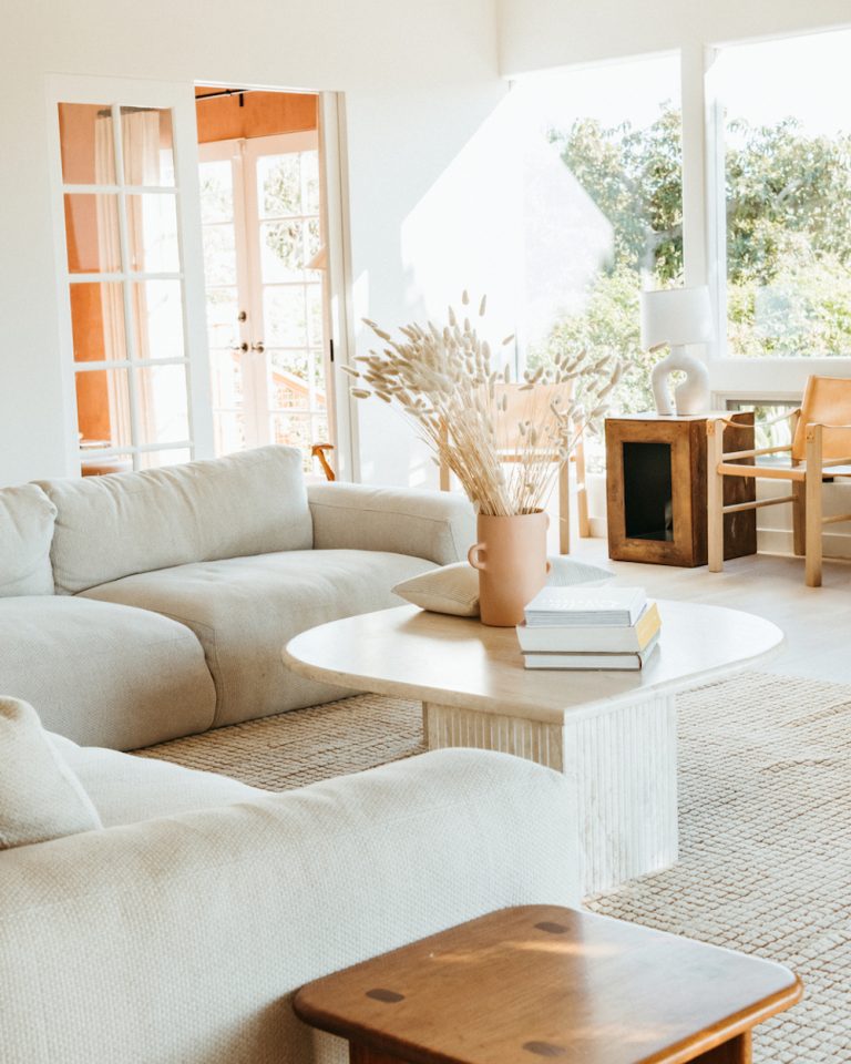 Airy, white living room with white walls, large light gray sectional couch, natural woven rug, and light wood coffee table with terra cotta vase.