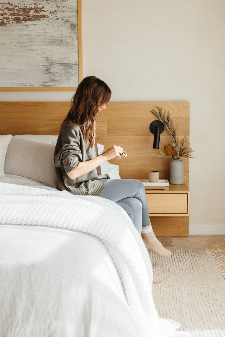 Brunette woman wearing loungewear sitting on bed in natural, minimalist bedroom lighting candle.