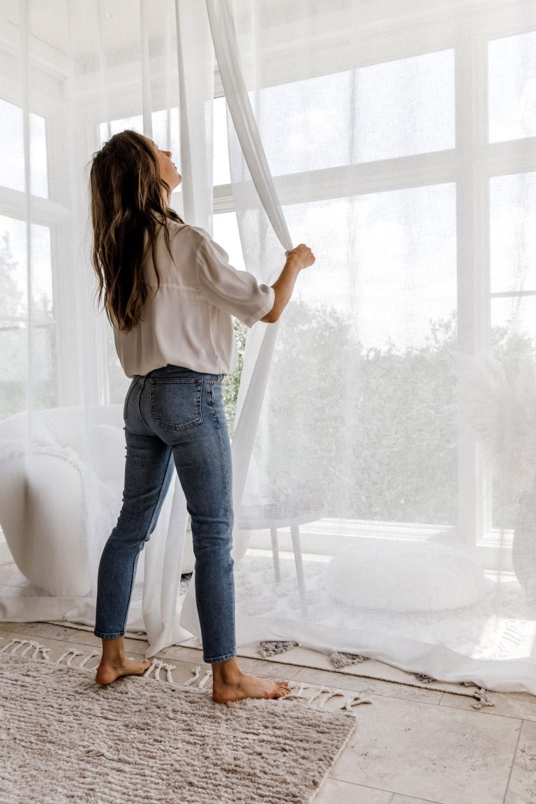 A brunette woman in a white button-down shirt and jeans moves white sheer curtains in a room.