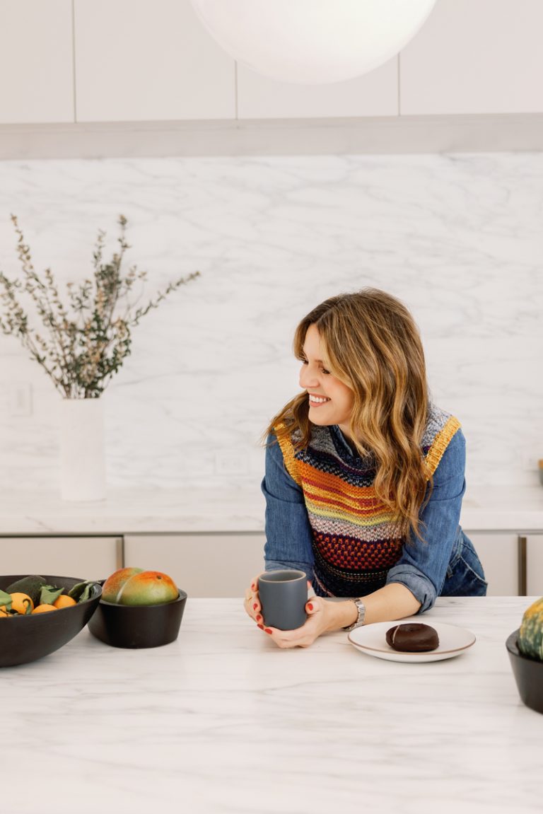 Candace Nelson holding coffee at marble kitchen counter surrounded by bowls of fruit and doughnut.
