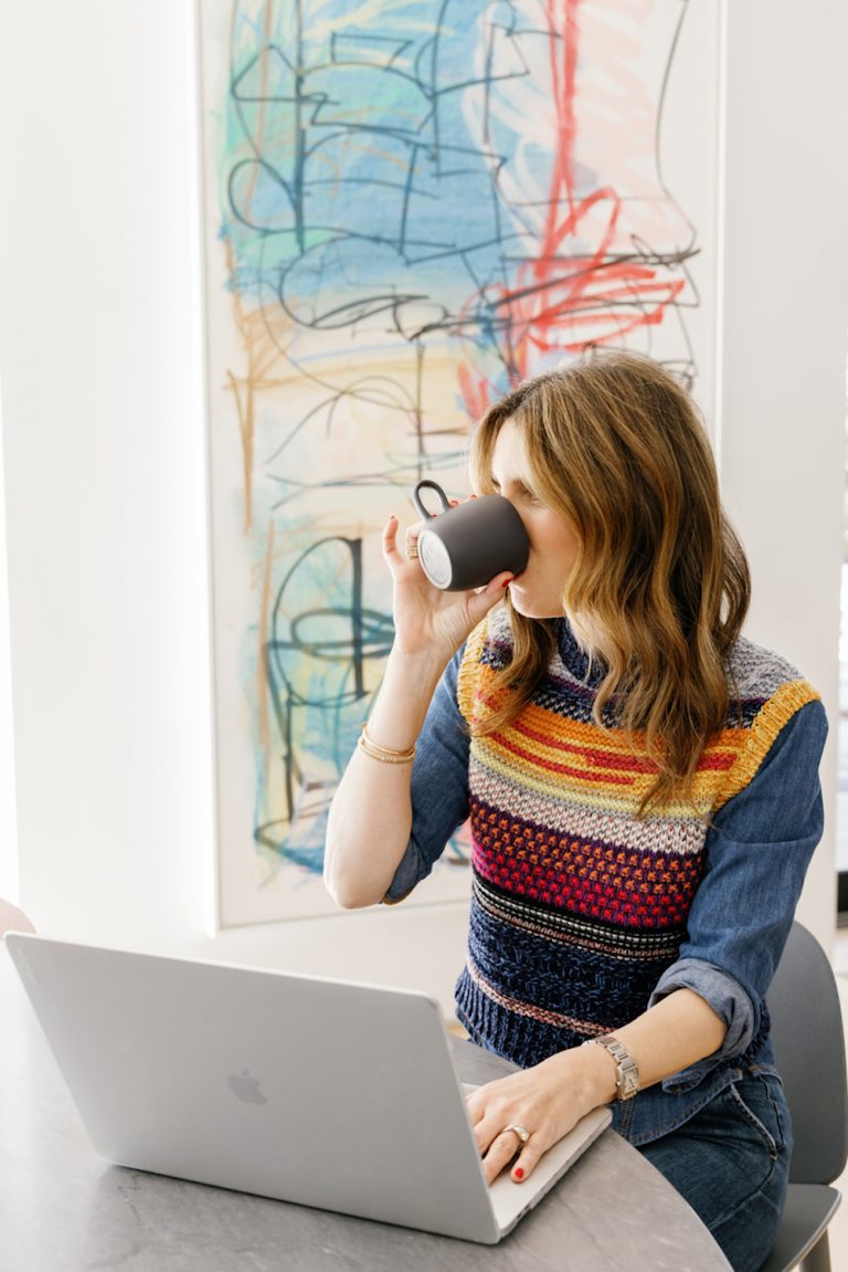 Brunette woman drinking coffee and working on laptop at table.