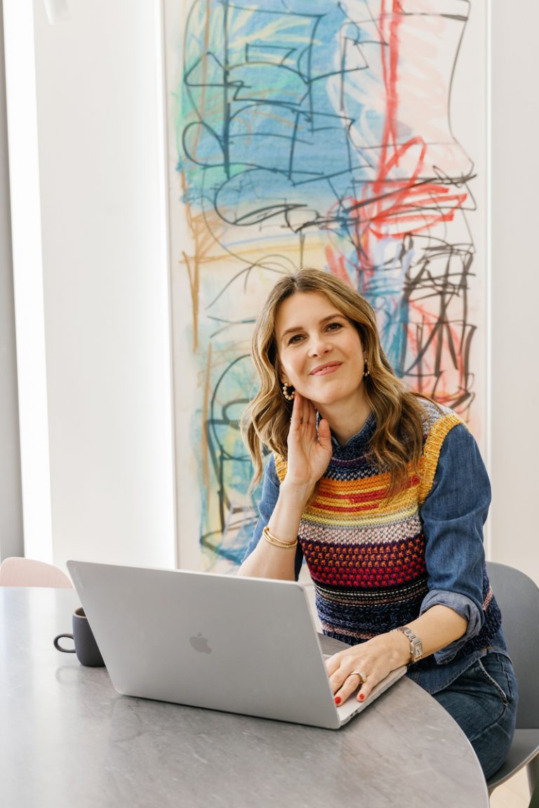 Candace Nelson wearing colorful sweater vest over denim shirt working at table on laptop.