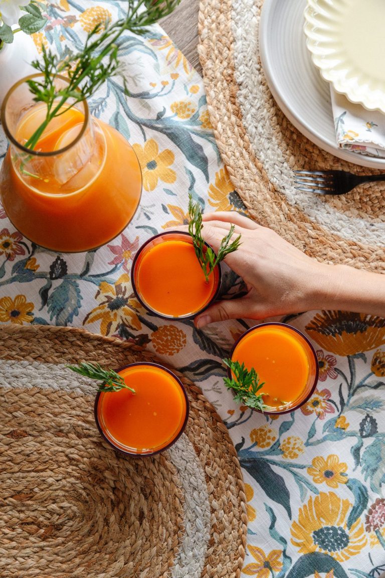 Three glasses of orange carrot juice on table with woven placemat and floral tablecloth.