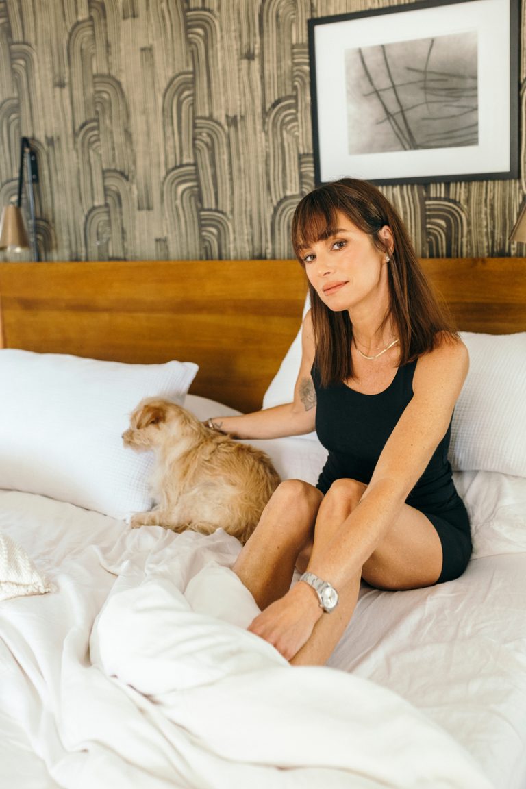 A brunette woman in a black tank top and shorts strokes a dog on her bed.