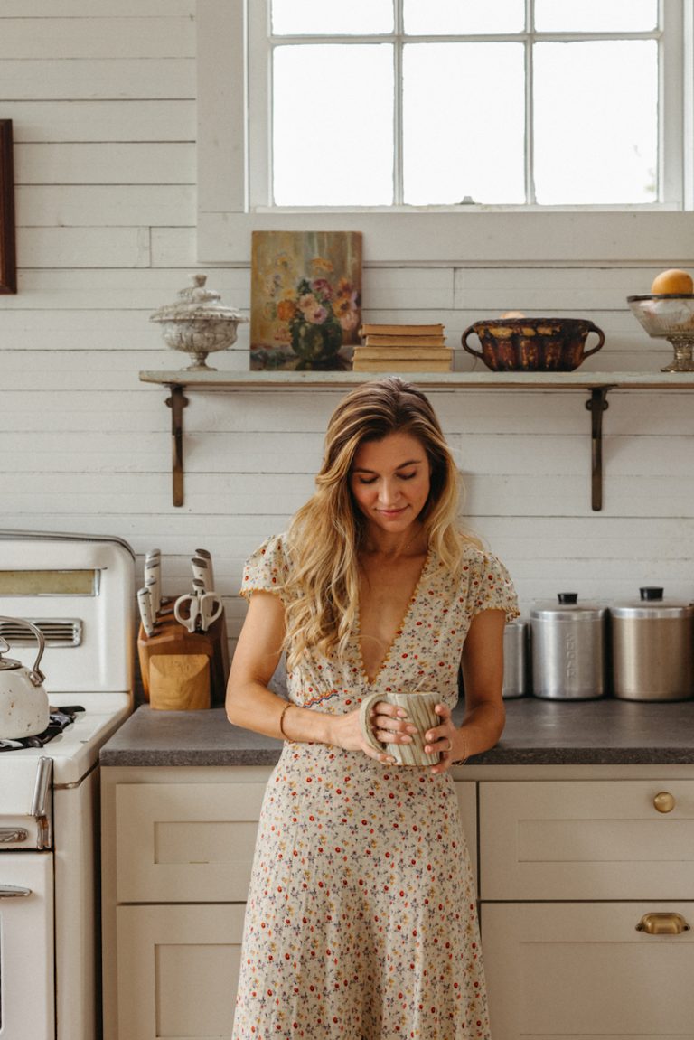 Claire Zinnecker wearing mid-length floral print dress holding cup of coffee in kitchen.