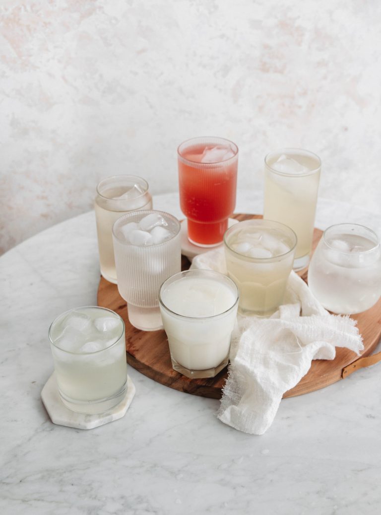 Various clear glasses of coconut water atop wood cutting board on marble table.