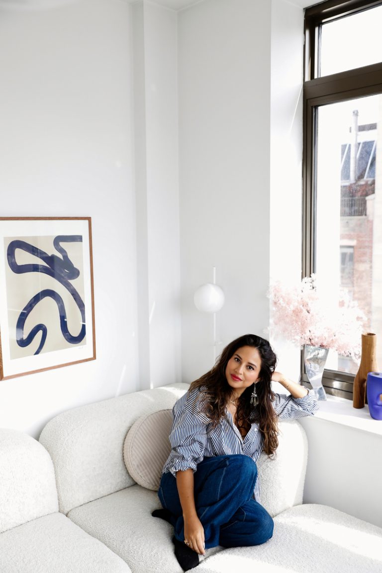 A brunette woman wearing a button down and jeans sitting on a cream sofa in a modern living room.