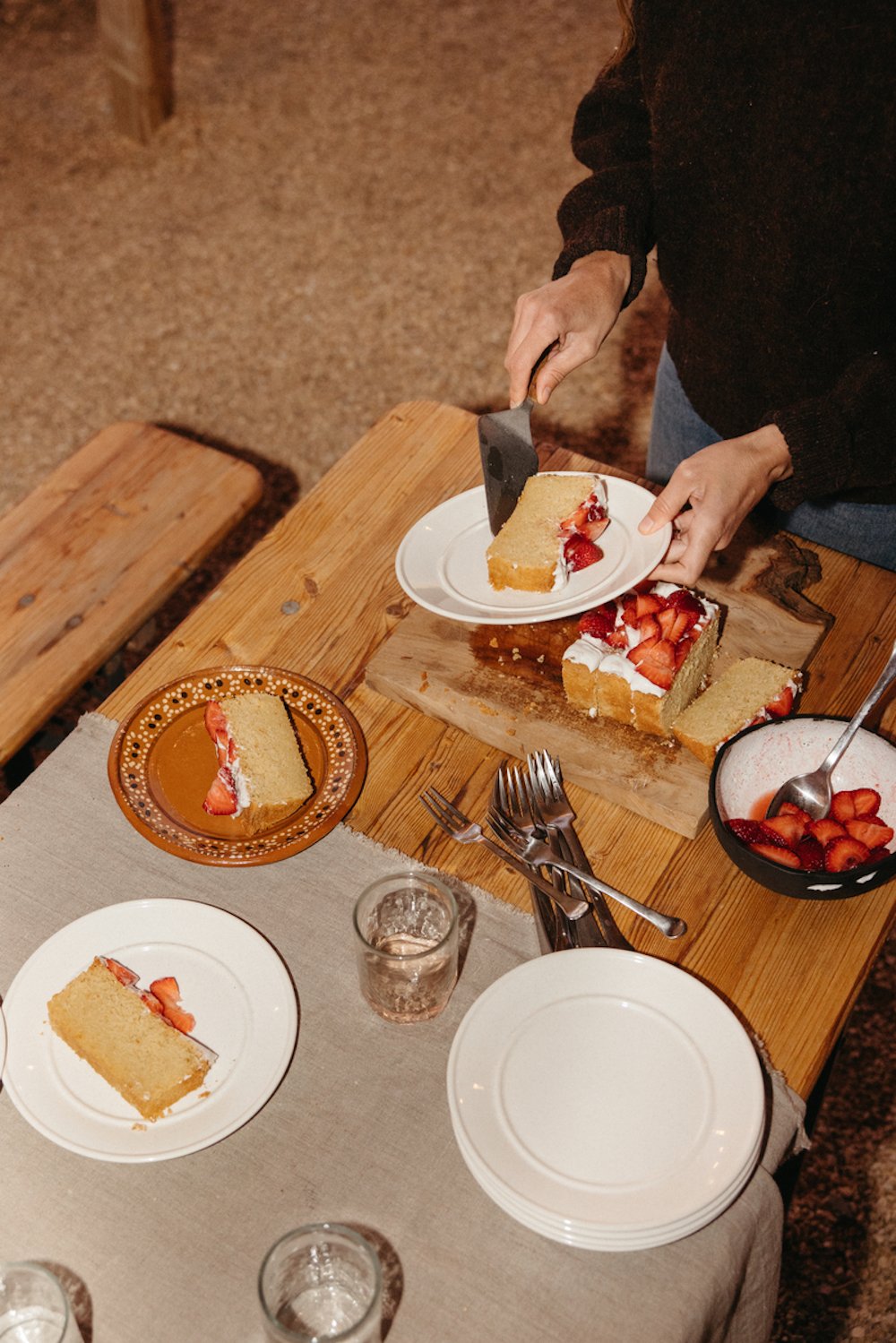 Woman serving slices of yellow loaf cake with strawberries onto various plates at long wooden table.