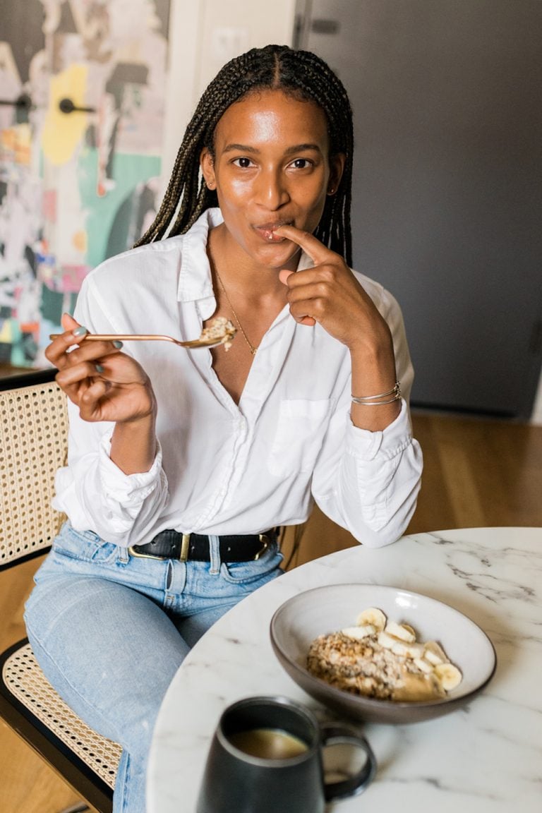 Woman wearing white button-down shirt and jeans eating banana oatmeal and drinking coffee at round marble table.
