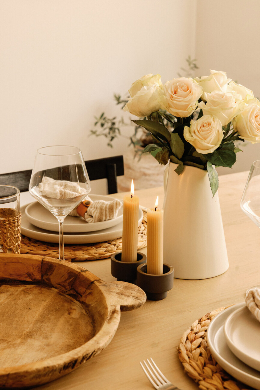 Cream roses in a white pitcher beside taper candles, woven placemats, pale plates, and a large wood bowl.
