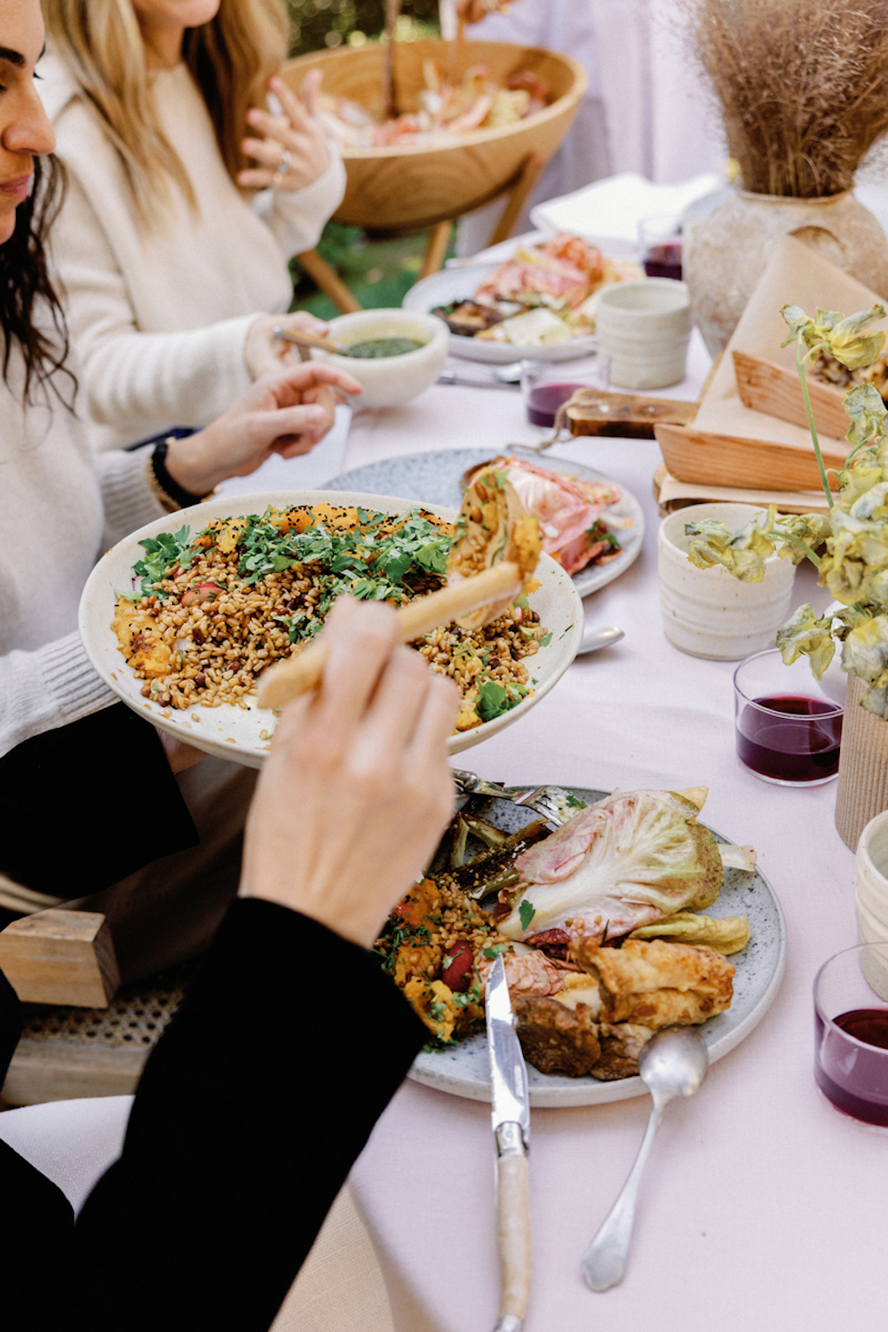 Women serving dishes family style at dinner table with pink tablecloth.