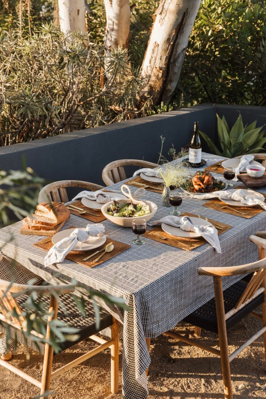Outdoor dining table with a patterned tablecloth, white place settings, and shared platters of food surrounded by greenery.