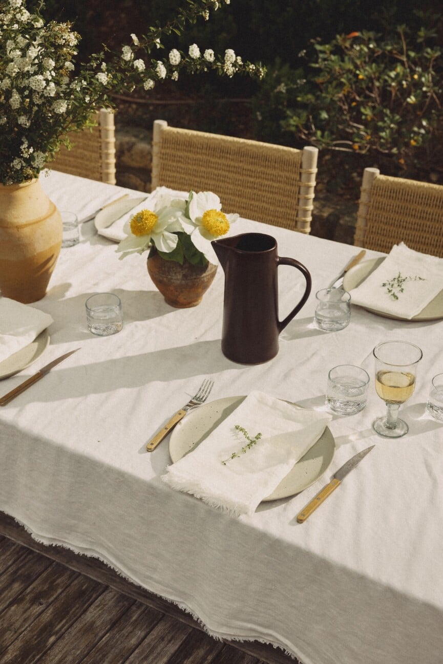 Outdoor table with white daffodils, a dark pitcher, linen napkins, and a leafy sprig at each place setting.