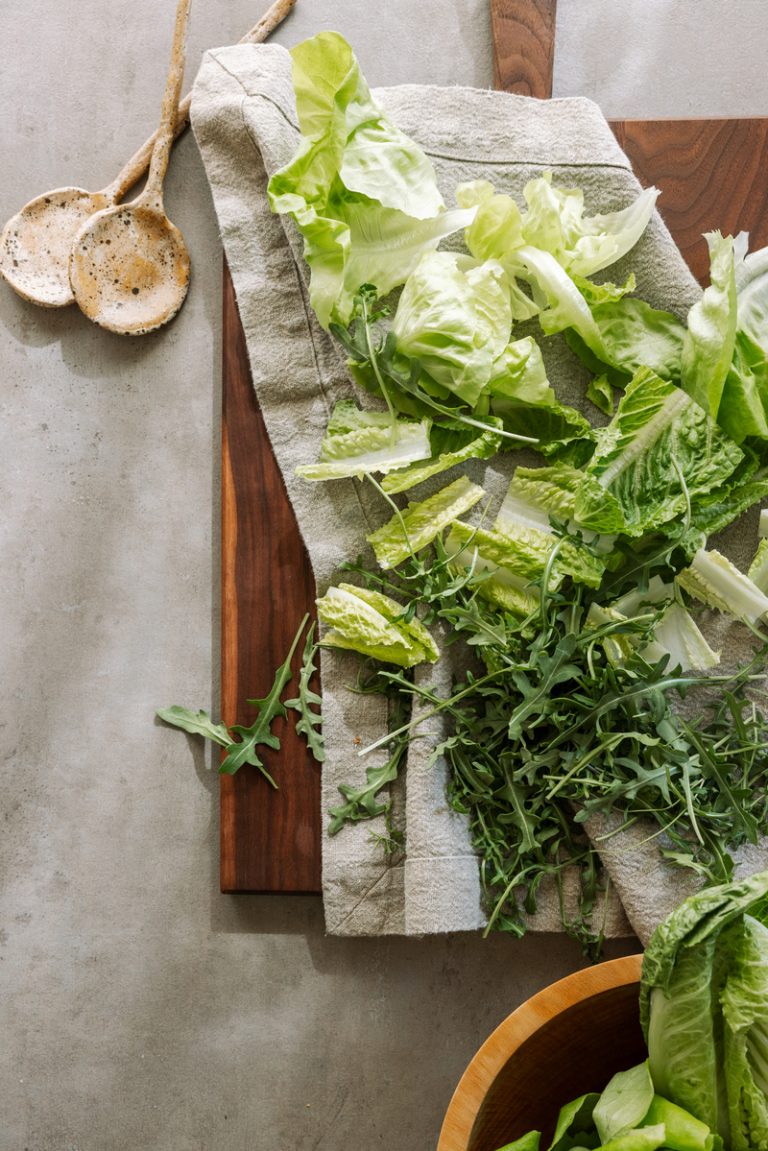 Green salad ingredients on maple wood cutting board with salad servers.