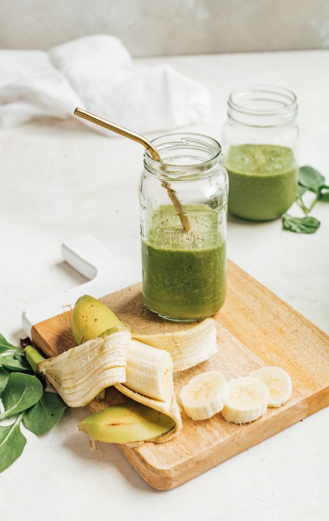Green smoothie with gold metal straw in Mason jar on wood cutting board with sliced banana and spinach