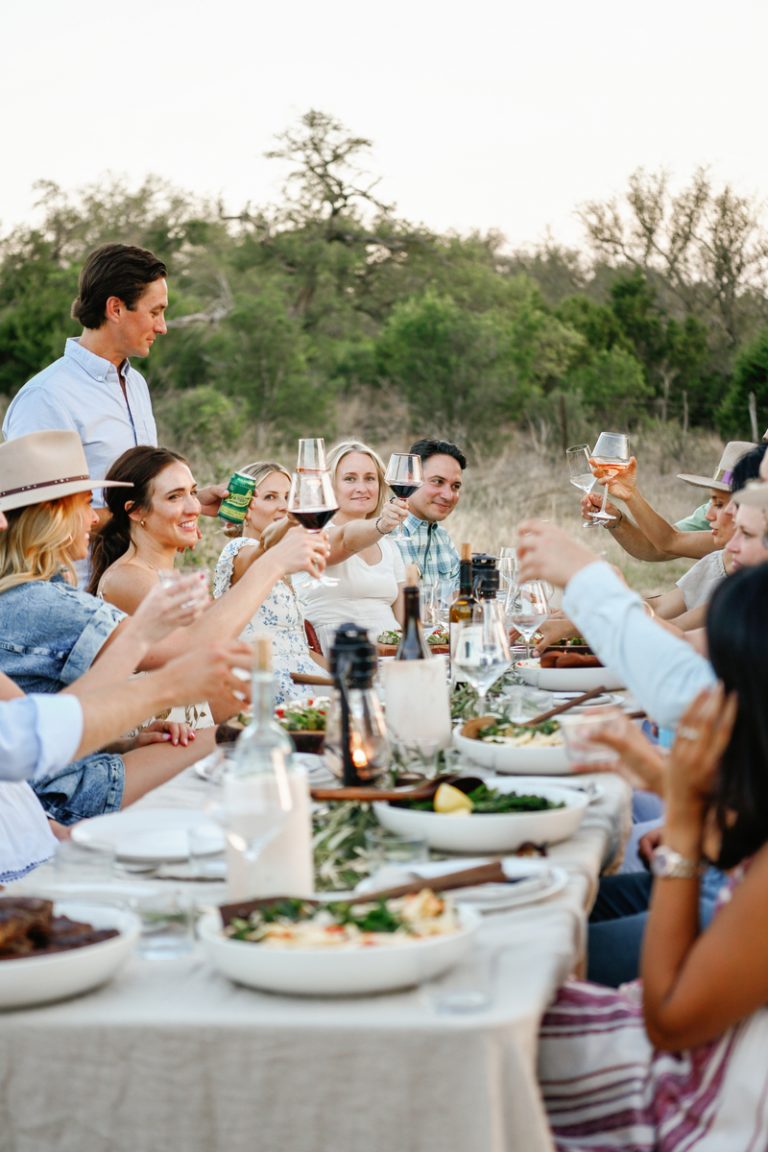 People raising wine glasses in cheers around long rectangular dining table outside.