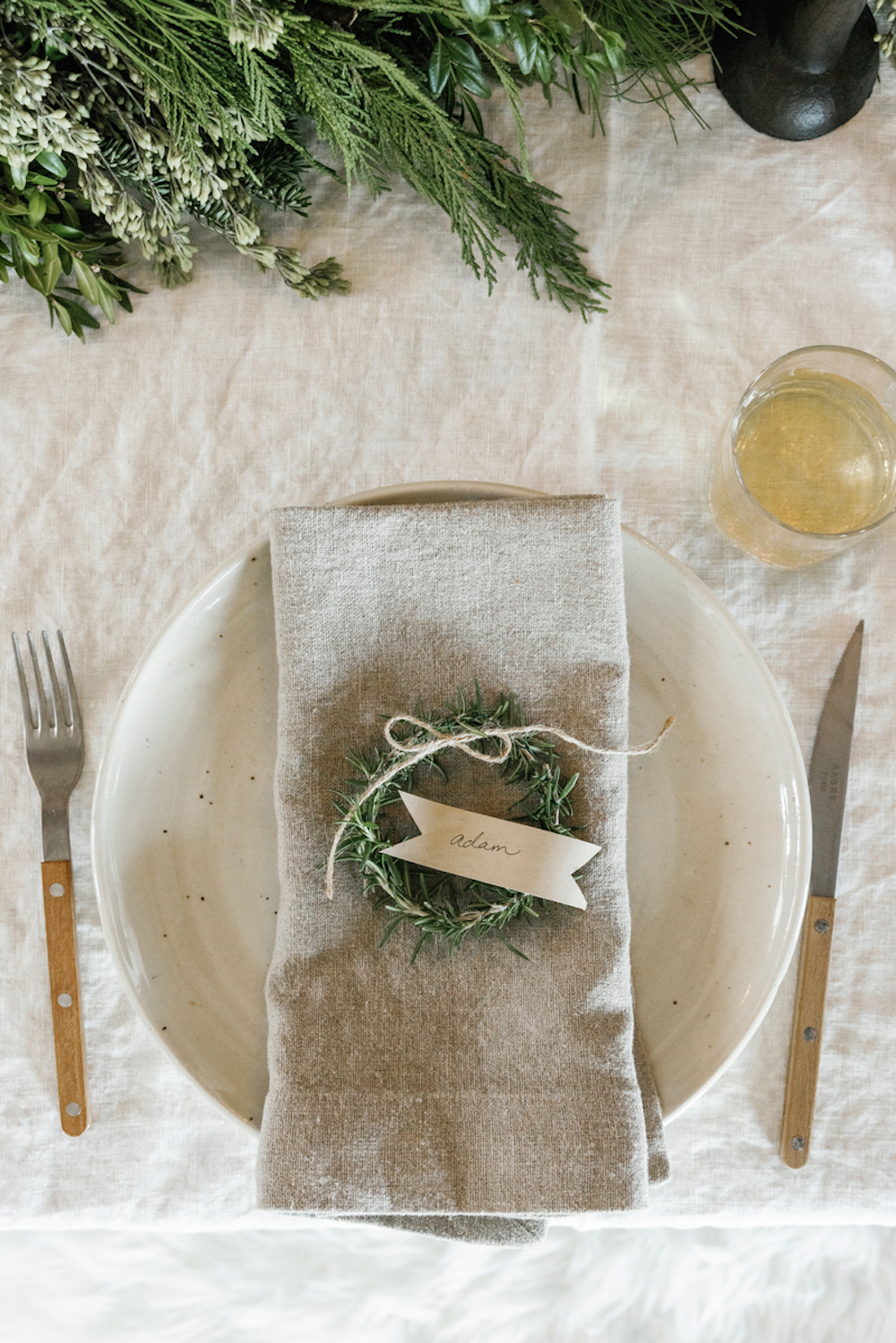 Simple holiday table setting with stoneware plate, wooden-handled silverware, clear wine glass, gray linen napkin, and rosemary wreath placecard.