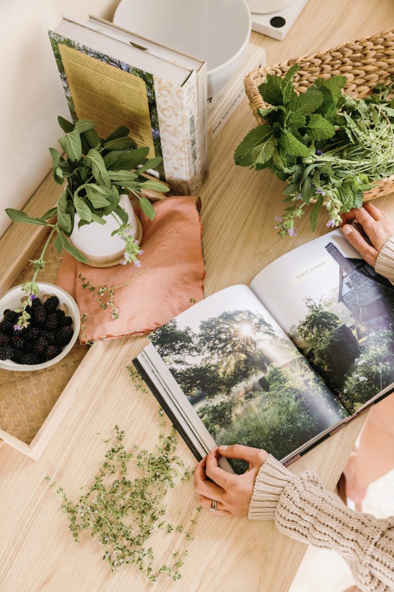 Woman reading book surrounded by indoor herbs in baskets and vases.