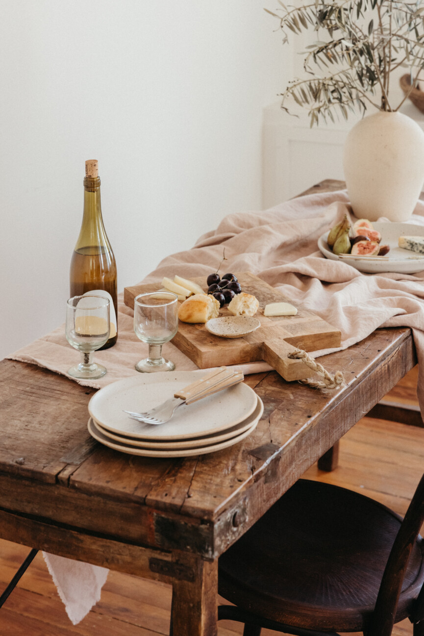 Rustic wood table with loosely draped blush linen, stacked ceramic plates, a cheese board, and branches in a white vase.