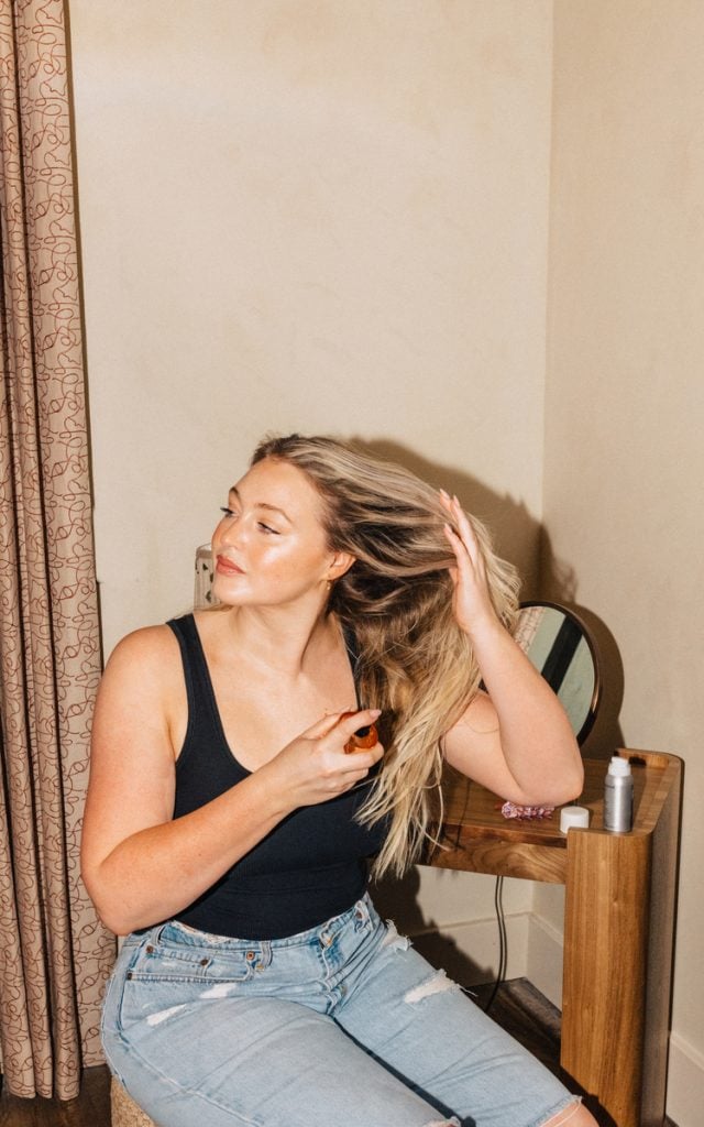 Blonde woman wearing black tank top and jeans sitting down spraying hairspray.