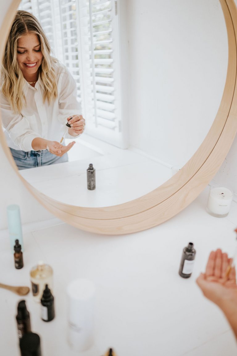 Blonde woman applying serum in bathroom mirror.