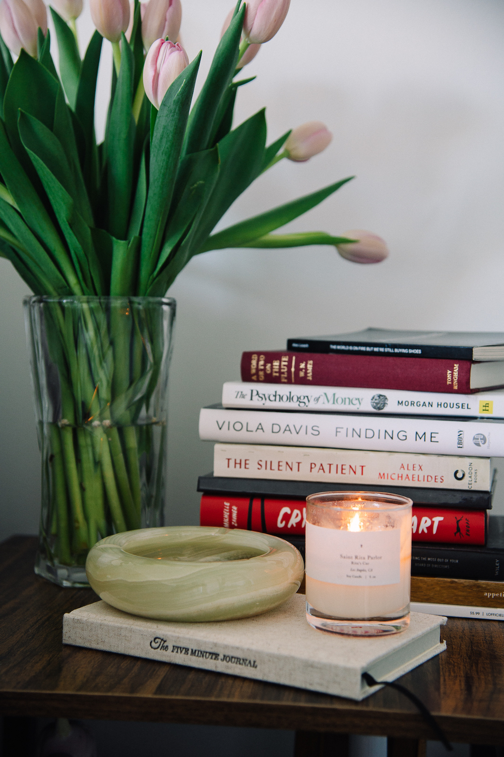 Stack of books, candle, and a bouquet of flowers on nightstand.