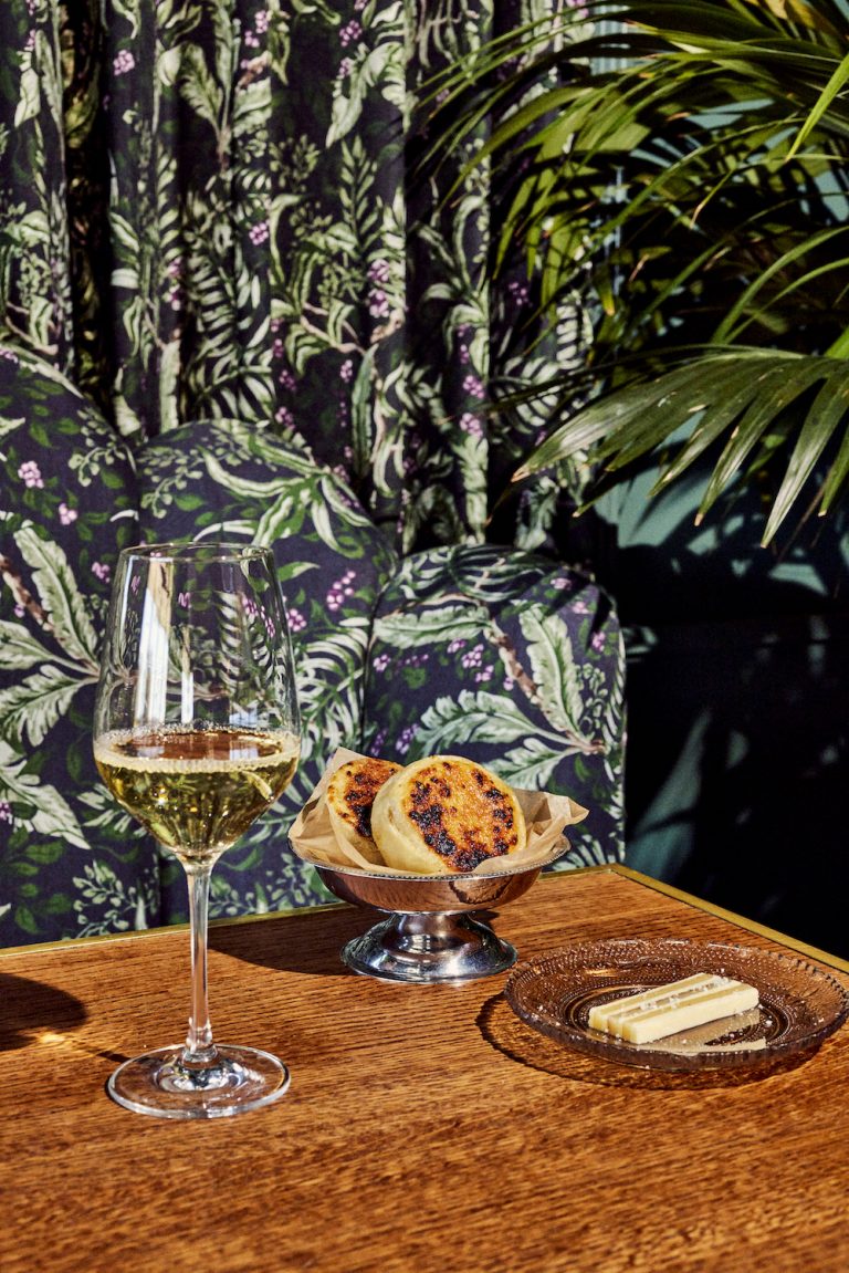 Copa de vino blanco, plato de plata con bollería y queso sobre una mesa de madera frente al papel pintado botánico.