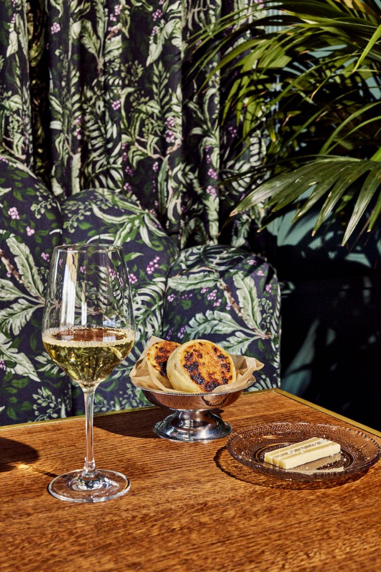 Copa de vino blanco, plato de plata con bollería y queso sobre una mesa de madera frente a un papel tapiz botánico.