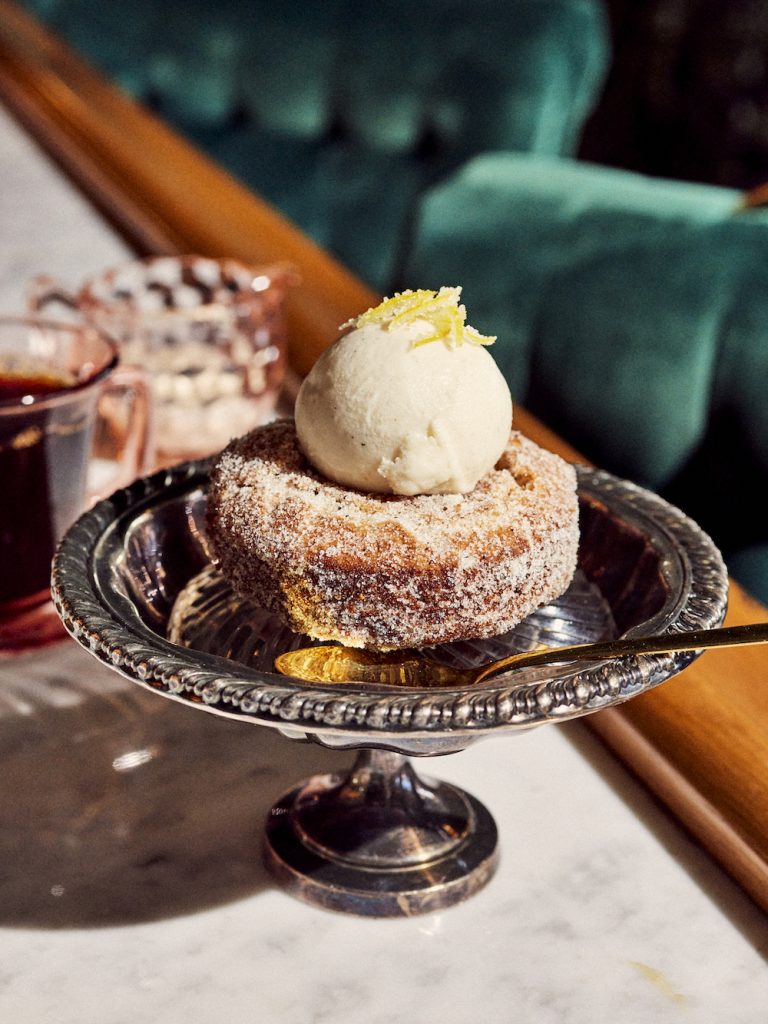 A donut topped with a scoop of vanilla ice cream on a silver plate with a gold spoon.