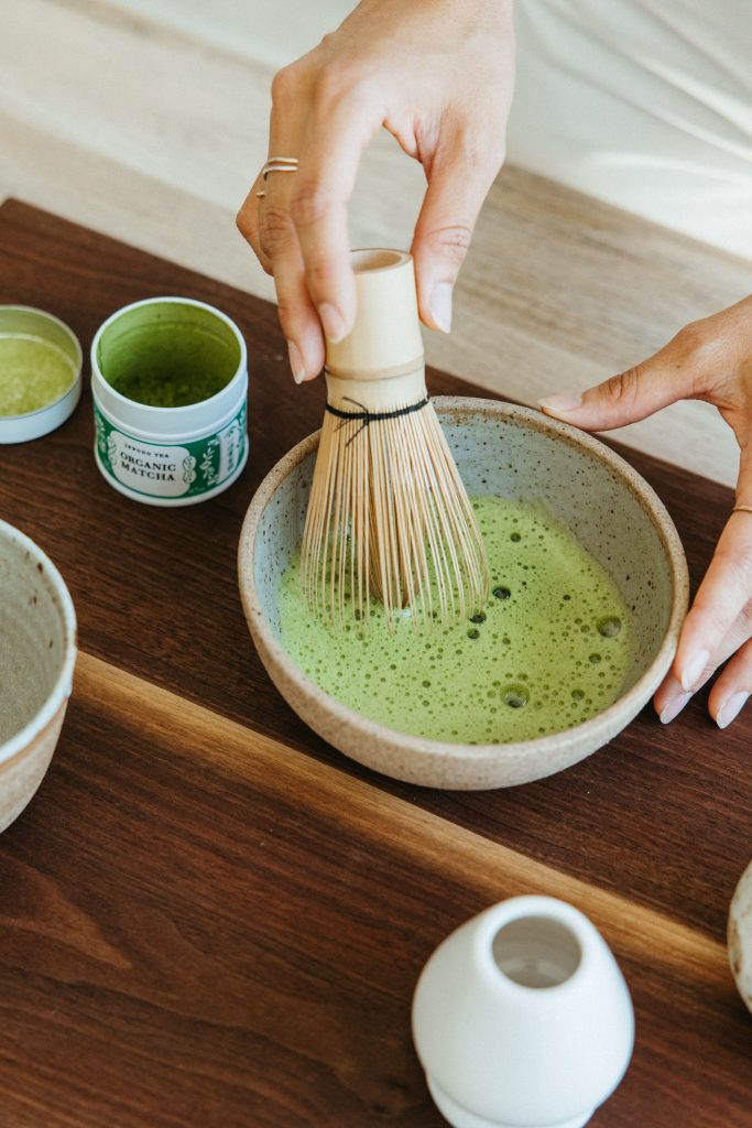 Making matcha with whisk is stoneware bowl on wooden cutting board.