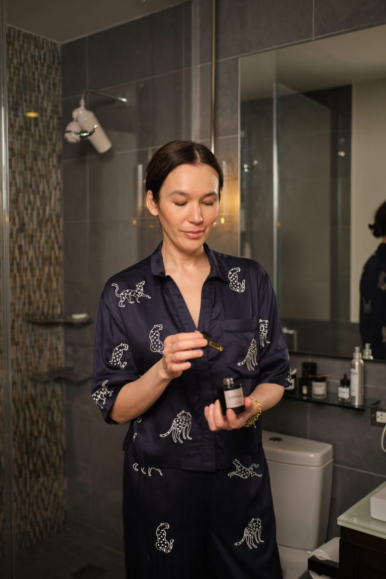 Brunette woman wearing silk blue pajamas applying skincare serum in bathroom.