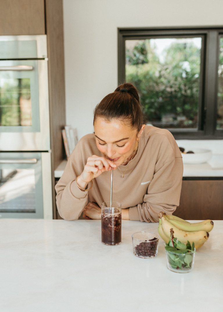 Brunette woman wearing light brown sweatshirt sipping blueberry, spinach, and banana smoothie in kitchen.