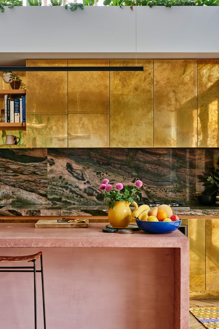 Cocina maximalista con gabinetes dorados, mosaicos abstractos y una isla de cocina rosa.