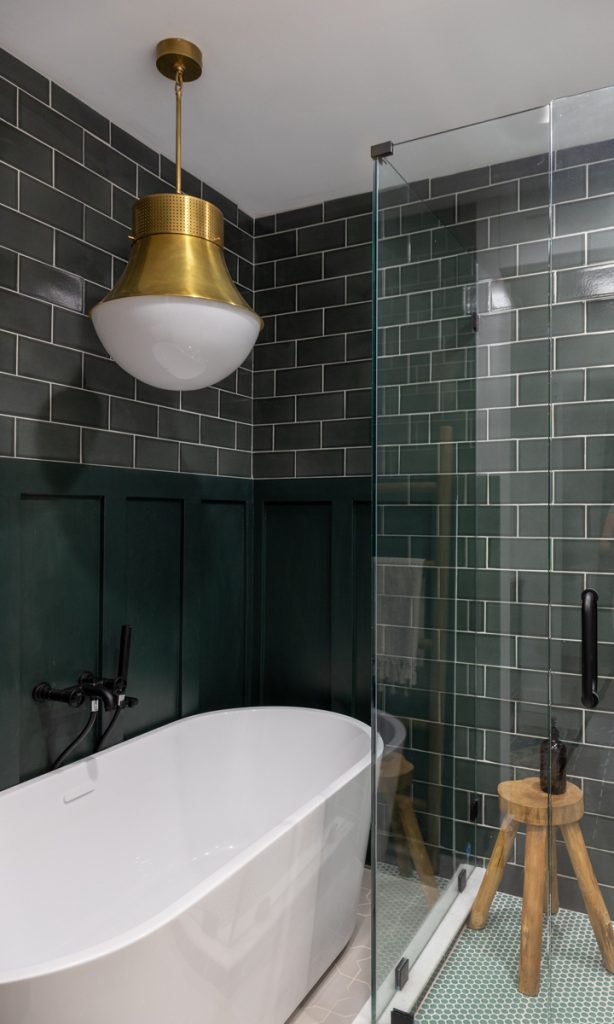 Dark green subway tiled bathroom with large white tub and pendant lighting.