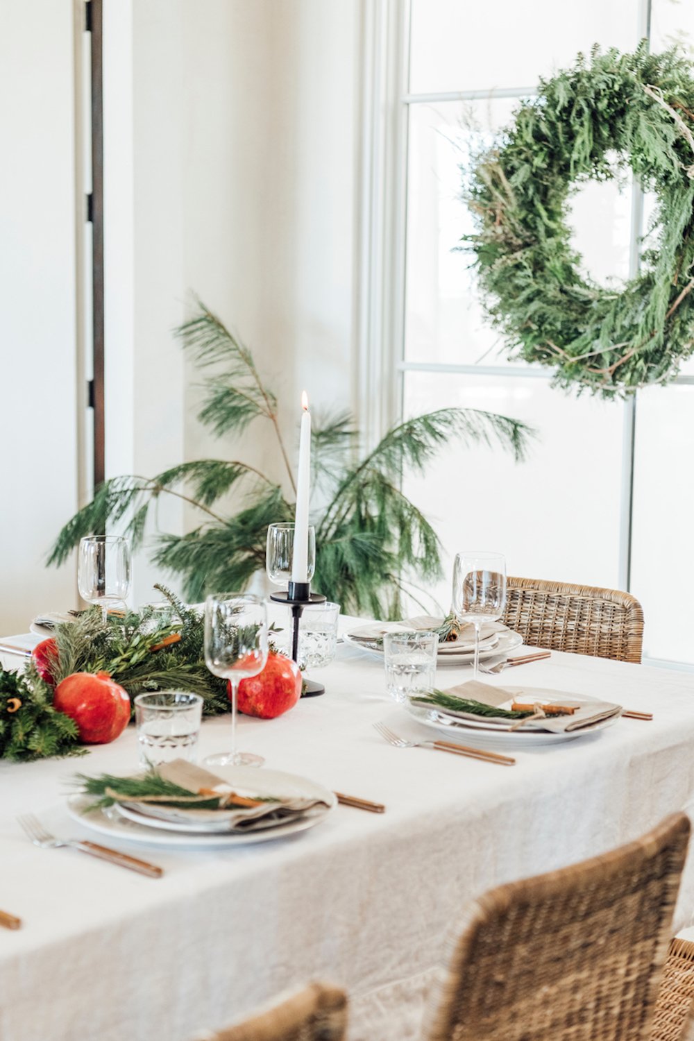 Modern holiday table setting with white tablecloth, modern drinking and wine glasses, white plates, gray linen napkins, and evergreen pine centerpiece with pomegranates.