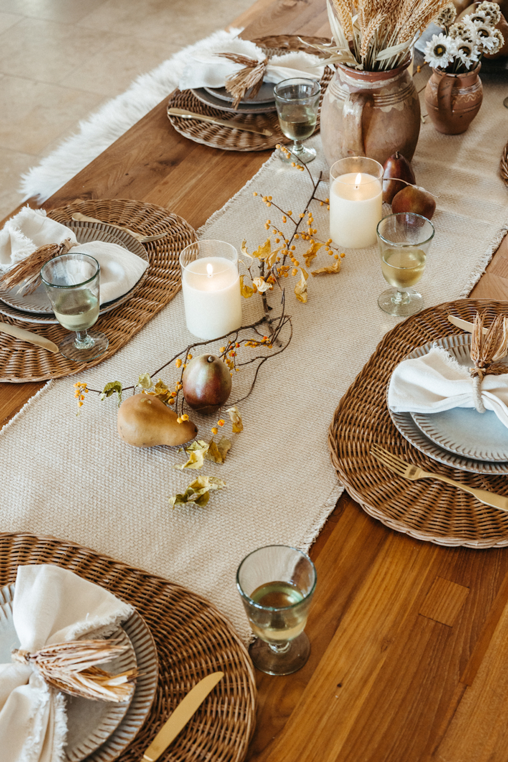 Modern Thanksgiving table setting with off-white textured table runner, white pillar candles, dried branches, and pears as a centerpiece, rattan chargers, and neutral dinnerware.