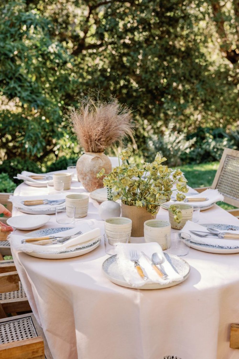 Natural outdoor table with light pink linen tablecloth, neutral dinnerware and flatware, and natural florals.